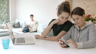 Sisters using smartphone together with Mom in background