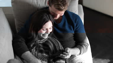Couple relaxing on sofa at home watching video on smartphone 