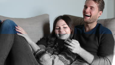 Couple relaxing on sofa at home watching video on smartphone 