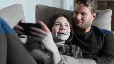 Couple relaxing on sofa at home watching video on smartphone 