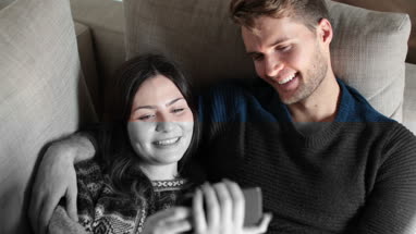 Couple relaxing on sofa at home watching video on smartphone 