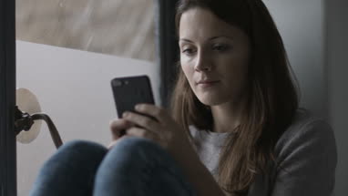 Young adult female sitting on window ledge with smartphone