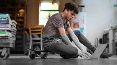 Young adult male sitting on skateboard using laptop