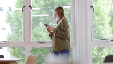 Businesswoman in office looking out of the window and at smartphone