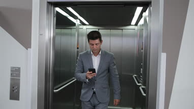 Businessman walking out of elevator in an office on smartphone