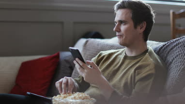 Young male using smartphone and streaming movie at home eating popcorn