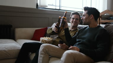 Young male couple relaxing on sofa with beer and popcorn