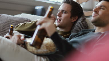 Young male couple relaxing on sofa with beer and popcorn