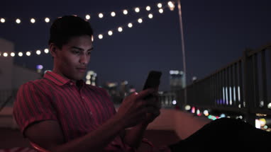 Young adult male looking at smartphone on a rooftop at night