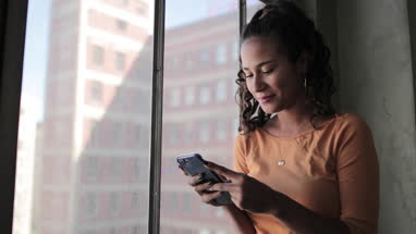 Young hispanic female looking out of window in loft apartment holding smartphone