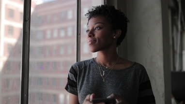 Young african american female looking out of window in loft apartment holding smartphone